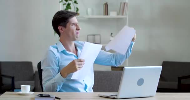 Mutlu erkek ofis çalışanı katip ofis masasında oturuyor, elinde belgeleri tutuyor, neşeyle dikkatsizce kağıt siparişleri dağıtıyor, kağıtları havaya dağıtıyor, iş yerinde oynamaktan keyif alıyor. — Stok video