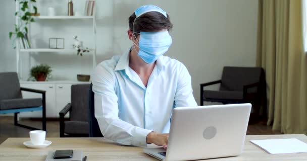 Funny office worker freelance joker se sienta en la mesa con el ordenador portátil, lleva tres máscaras médicas protectoras, cerró los ojos y la boca de la nariz, escondió la cara del virus, tipos a ciegas, al azar toca la taza de café — Vídeo de stock