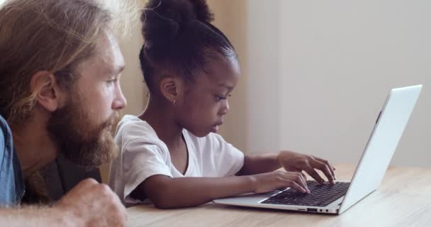 Γενειοφόρος Καυκάσιος πατέρας βοηθήσει biracial μικρή κόρη με την εργασία στο σπίτι στο φορητό υπολογιστή, Ευρωπαίος μπαμπάς και μικρό κορίτσι της Αφρικής μελέτη παιδί μαζί για την απομόνωση καραντίνας, e-learning έννοια — Αρχείο Βίντεο