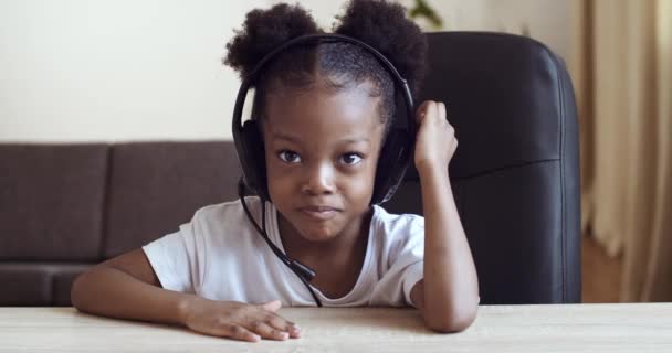 Retrato da menina pré-escolar africana ativa bonito sentado à mesa em casa, usando fones de ouvido falando no microfone fone fone fone de ouvido se comunicando online. Afro-americana criança étnica pequena senhora gestos — Vídeo de Stock