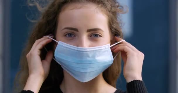 Porträt eines jungen Mädchens Teenager lockig schöne Frau setzt medizinische chirurgische Schutzmaske auf weibliche Gesicht, steht im Freien allein isoliert in die Kamera mit blauen Augen, Virus-Pandemie Zeit — Stockvideo