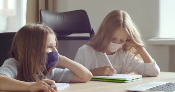To skolejenter i beskyttelsesmasker som sitter sammen ved bordet hjemme i klasserommet, ser på laptop-lerretet, ler og gjør lekser på nettet, konseptet utdanning – stockvideo
