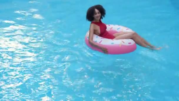 Samotná mladá africká žena v červených plavkách plavala, tahala, veslovala v bazénu v nafukovacím kruhu růžové koblihy a smála se na kameru. Etnicity Afro žena relaxovat v modré vodě s gumové kroužkové trubice — Stock video