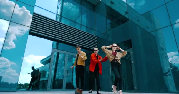 Drie jonge tieners in stijlvolle heldere shirts en een zonnebril poseren lopen in beweging voor de camera, bewegen sierlijk, vertonen modieuze kleding, twee jongens met meisje dansen op straat — Stockvideo