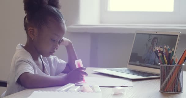 Roztomilá školačka kreslí na papíře, jak sedí za stolem a dívá se na monitor notebooku. Africké americké etnické dítě sledující video online mluvit s učitelem vzdáleně přes webkameru, vychovatel odpovědí, e-learning — Stock video