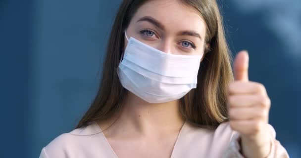 Jonge zakenvrouw geïsoleerd buiten draagt beschermende medische chirurgische masker op haar gezicht tonen duimen omhoog gebaar van goedkeuring, meisje student drukt steun ja teken symbool van tevredenheid — Stockvideo