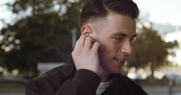 Retrato de hombre joven morena en ropa elegante se para solo al aire libre, lleva auriculares inalámbricos, habla por teléfono, escucha al interlocutor, responde a la llamada, se comunica de forma remota, sonriendo de cerca — Vídeo de stock