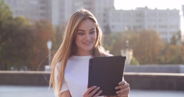 Zblízka portrét šťastné mladé ženy stojící venku, aby video chat na digitální tablet mluvit ve webové kameře. Krásná kavkazská radostná žena nosí sluchátka mluvící na videohovor na zařízení on-line — Stock video