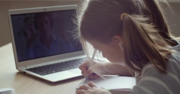 Se fra bak skolejentebarn som sitter hjemme i karantene isolasjon, ser på laptop-skjermkonferanse med læreren, bruker hodetelefoner lytter til leksjoner, utdanningskunnskap fjernstyrt – stockvideo