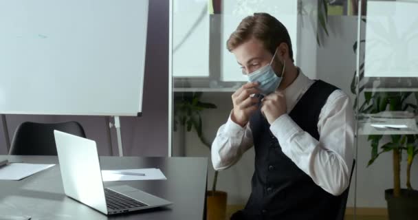 Zakenman freelancer in gezicht medisch masker zit aan tafel berichten online op Covid-19 lockdown. Blanke man typt op laptop toetsenbord. Manager werkt op afstand van kantoor in coronavirus pandemie — Stockvideo