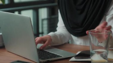 İşkadını tesettüre odaklanmış müslüman iş kadını internet servis laptopuyla çalışan kız çocuğu çalışma ortamındaki içme suyunu bir bardak içecekle birlikte kullanıyor.