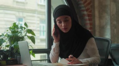 Stresli, çok çalışan, çok görevli, Müslüman iş kadını, tesettürlü, kadın, tesettürlü, iş kadını, konuşan, cep telefonlu, dizüstü bilgisayara bakan, not defterinde veri arayan, iş sıkıntısı çeken.