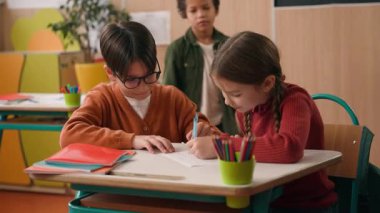 Sınıfta üç farklı çocuk var. İki beyaz sınıf arkadaşı ve kız birlikte resim çiziyorlar. Afro-Amerikan çocuk resme bakıyor.