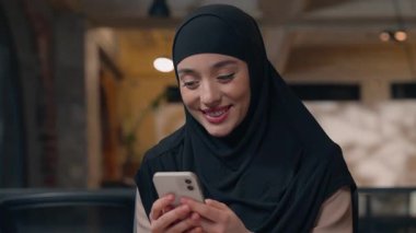 İş yerinde çalışan Müslüman iş kadını tesettürlü cep telefonu işkadınıyla sohbet ederken gülümsüyor ofis kızı Arap kadın çalışan akıllı telefon uygulamasını kullanarak internetten SMS yazarken mutlu oluyor.