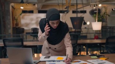 Telefonla konuşan Müslüman iş kadını kağıtları analiz ediyor iş kadını ofiste çoklu görev ile meşgul cevap ver proje istatistiklerini tartış tesettürlü kadın Arap kız aşırı çalışma görevlerini yönet