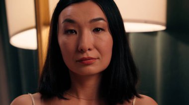 Düşünceli, sakin, ciddi, yetişkin, etnik, Asyalı, Çinli bir kadın. Japon bayan, akşam evde kameranın güzelliğine, cilt sağlığına bakıyor.