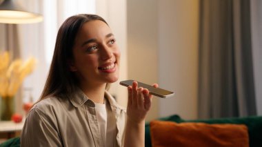 Beyaz, mutlu, gülümseyen kadın, cep telefonu uygulamaları kullanan, sesli mesaj kaydeden, evdeki akıllı telefondan ses tanıma teknolojisi gönderen, cep telefonu kullanan.