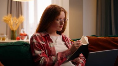 Yoğunlaşmış Kafkas kadın kız ev öğrencisi serbest yazar not defterine notlar yazar bilgi arayan dizüstü bilgisayarı kullanarak evde e-öğrenim yapar.