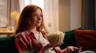 Oturma odasındaki Kafkasyalı mutlu kadın dinlenme haftasonu kanepede dinleniyor genç kız molası veriyor evde oturmuş patlamış mısır yiyor TV 'de film serisi seyrediyor.