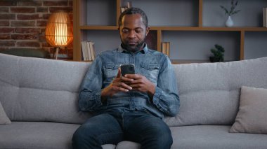 Mutlu Afrikalı Amerikalı bir adam telefoncu bir erkeği evde cep telefonuyla koltukta dinlenirken cep telefonuyla gezerken, sohbet ederken internetten alışveriş yaparken internet uygulaması e-ticaretinde eğleniyor.