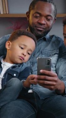 Mutlu Afro-Amerikan ailesi evde cep telefonu uygulaması kullanıyor. Babaları akıllı telefon aletlerine bakıyor. Gülümsüyor, sosyal medya çocuklarıyla konuşuyor.