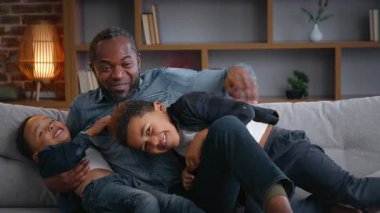 Mutlu Afro-Amerikan ailesi kanepede oynaşıyor. Babaları iki küçük çocuğu gıdıklıyor. Çocuklar gülüyor.