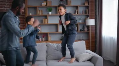 Çocuklarla dans eden pozitif Afro-Amerikan ailesi hareketli müzik dinleyen baba iki çocuklu baba çocuklar oturma odasında dans ediyor kanepeye zıplıyor hafta sonu taşınma kredisiyle eğleniyorlar.