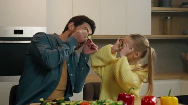 Mutfaktaki mutlu beyaz aile oyun oynuyor, eğleniyor, kafkasyalı küçük kız çocuk babası, elinde salatalık dilimleri, gözlerinde gözlük, sebze halkaları ve eğlenceli yemekler tutan çocuk.