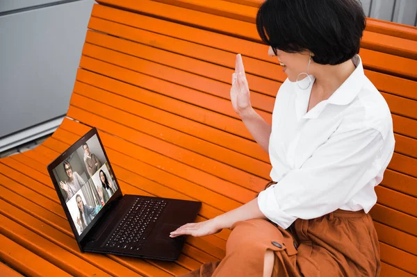 Video conference. Caucasian attractive girl with eyeglasses dressed in a formal clothes greets her colleagues communicating with them via video conference using laptop, while sitting on a bench outdoors