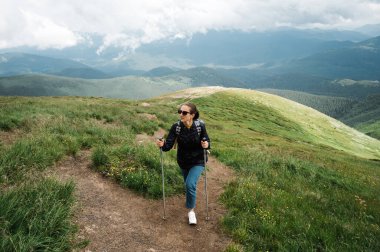 Yürüyüş sopalarıyla dağın tepesine tırmanan genç bir kadın yürüyüş kıyafetleriyle ve etrafındaki dağların muhteşem manzaralarıyla eğleniyor.