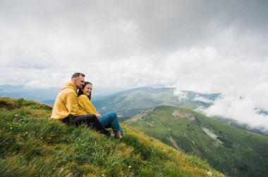 Dağlarda yürüyüş. Birbirine aşık çift dağın tepesine tırmandı, çimenlerin üzerinde otururken dinleniyorlar, birbirlerine sarılıyorlar ve eğleniyorlar ve ilham verici dağ manzarası