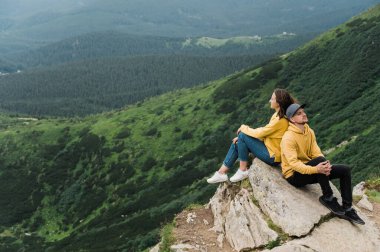 İlham verici bir manzara. Dağlarda aşk. Aşık mutlu çift uçurumun kenarında oturur. Arka planda muhteşem bir dağ manzarası olan büyük taşın üstünde sırt sırta.