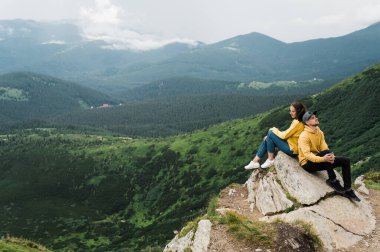 İlham verici bir manzara. Dağlarda aşk. Aşık mutlu çift uçurumun kenarında oturur. Arka planda muhteşem bir dağ manzarası olan büyük taşın üstünde sırt sırta.