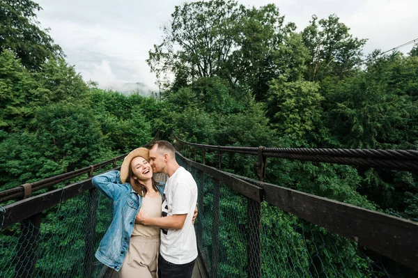 Mutlu ve neşeli bir çift, asma köprüde dikilirken birbirine sarılıyor. Güzel doğayla çevrili.