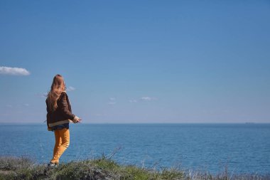 Sarı kot pantolon ve koyun derisi ceketli sarı uzun saçlı bir kız güneşli bir bahar gününde, denizüzerinde bir uçurum boyunca bir yol boyunca yürüyor.