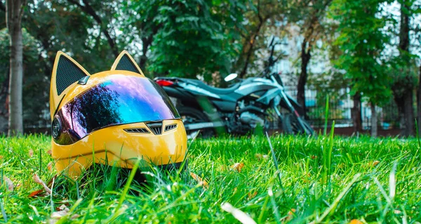 El casco amarillo de una chica motociclista de una motocicleta con espejo de vidrio y orejas se encuentra en la hierba verde sobre el fondo de una motocicleta gris en otoño . — Foto de Stock