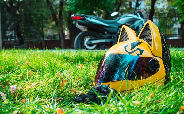 Helm kuning seorang gadis pengendara sepeda motor dengan kaca cermin dan telinga terletak di rumput hijau dengan latar belakang sepeda motor abu-abu di musim gugur . — Stok Foto