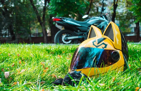 Helm kuning seorang gadis pengendara sepeda motor dengan kaca cermin dan telinga terletak di rumput hijau dengan latar belakang sepeda motor abu-abu di musim gugur . — Stok Foto