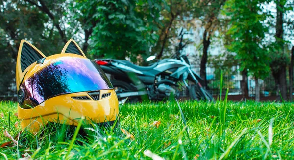 El casco amarillo de una chica motociclista de una motocicleta con espejo de vidrio y orejas se encuentra en la hierba verde sobre el fondo de una motocicleta gris en otoño . — Foto de Stock