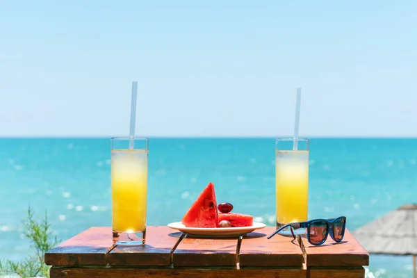 Two glasses of fresh tropical fruit juice on the beach, and a plate with grapes and watermelon on the table, against the sea. Holiday and vacation concept