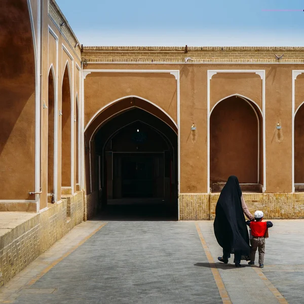 Une femme non identifiable portant une robe noire traditionnelle en Iran, connue sous le nom de chador, tient la main d'un jeune garçon. Architecture islamique en arrière-plan avec des arches antiques — Photo