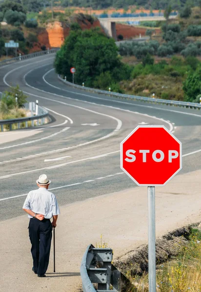 Homem idoso caminhando com uma bengala passando por um sinal de parada vermelho que leva a uma estrada curva - imagem conceitual — Fotografia de Stock