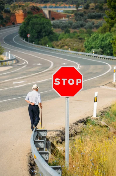 Homem idoso caminhando com uma bengala passando por um sinal de parada vermelho que leva a uma estrada curva - imagem conceitual — Fotografia de Stock