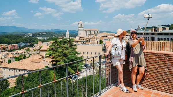 Två kvinnliga turist vänner ta en selfie med en selfie-stick med utsikt över en Flygfoto över den gamla staden Girona inklusive katedralen landmärken — Stockfoto