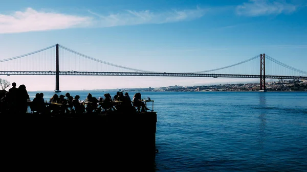 Silhuetten av människor avkopplande utomhus restaurang terrassen med utsikt över den ikoniska 25 April-bron i Lissabon, Portugal — Stockfoto