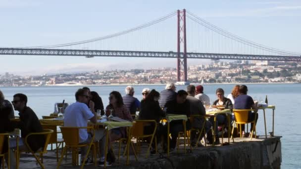 Die menschen entspannen sich auf der terrasse des restaurants mit blick auf die ikonische brücke 25 april in lissbon, portugal — Stockvideo