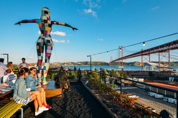 Vista del puente del 25 de abril con personas sentadas en la terraza del bar de LX Factory en Alcántara durante la puesta de sol en la ciudad de Lisboa, Portugal — Foto de Stock