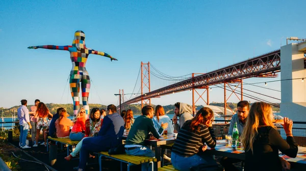 在葡萄牙里斯本市日落时分,人们坐在阿尔坎塔拉Lx工厂的酒吧露台上观看4月25日桥的景色 — 图库照片