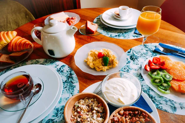 Mesa de desayuno con una variedad de alimentos que incluyen cereales, yogur, huevos revueltos, fruta, croissant y bebidas como té, café y jugo de naranja. — Foto de Stock