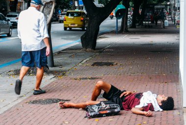 Ipanema, Rio de Janeiro, Brezilya zengin mahallede yoğun yaya kaldırımda yayalar onun yanında yürürken evsiz genç adam kaba uyku